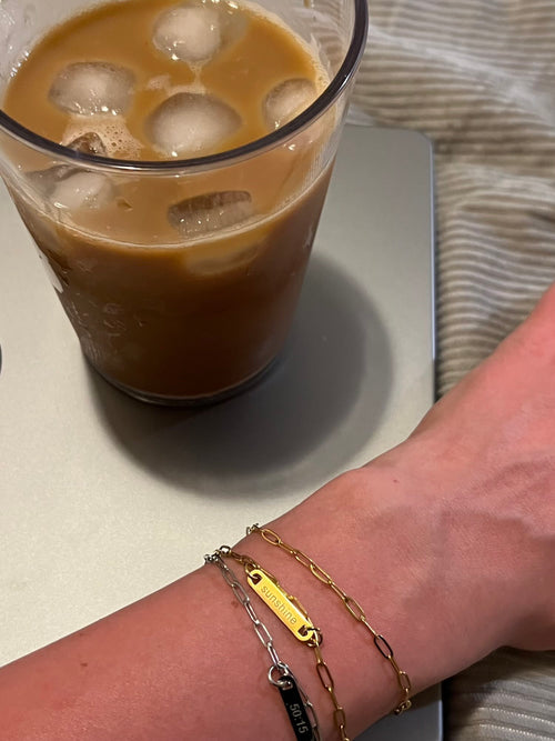 Two bracelets on a wrist next to an iced coffee sitting on top of a laptop on soft surface. Bracelets are paperclip chain with an engraved charm. Model wearing one gold and one silver engraved and an extra chain of gold for style- each sold separately.