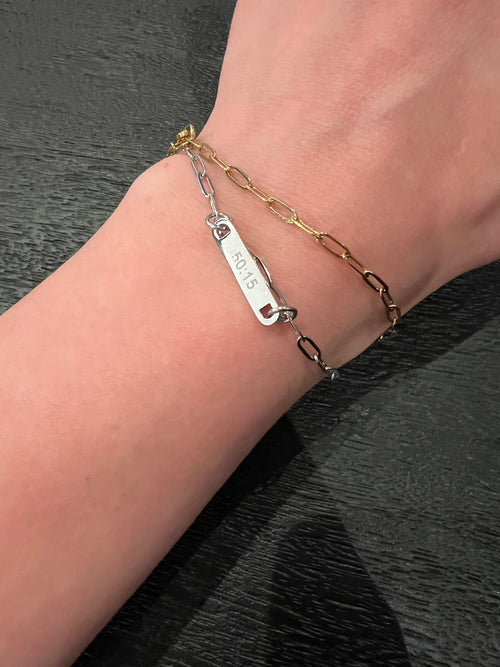 Two bracelets on a wrist against a dark background. A silver chain showing an engraving "50:15" and the gold only showing chain. Model wearing one gold and one silver- each sold separately.