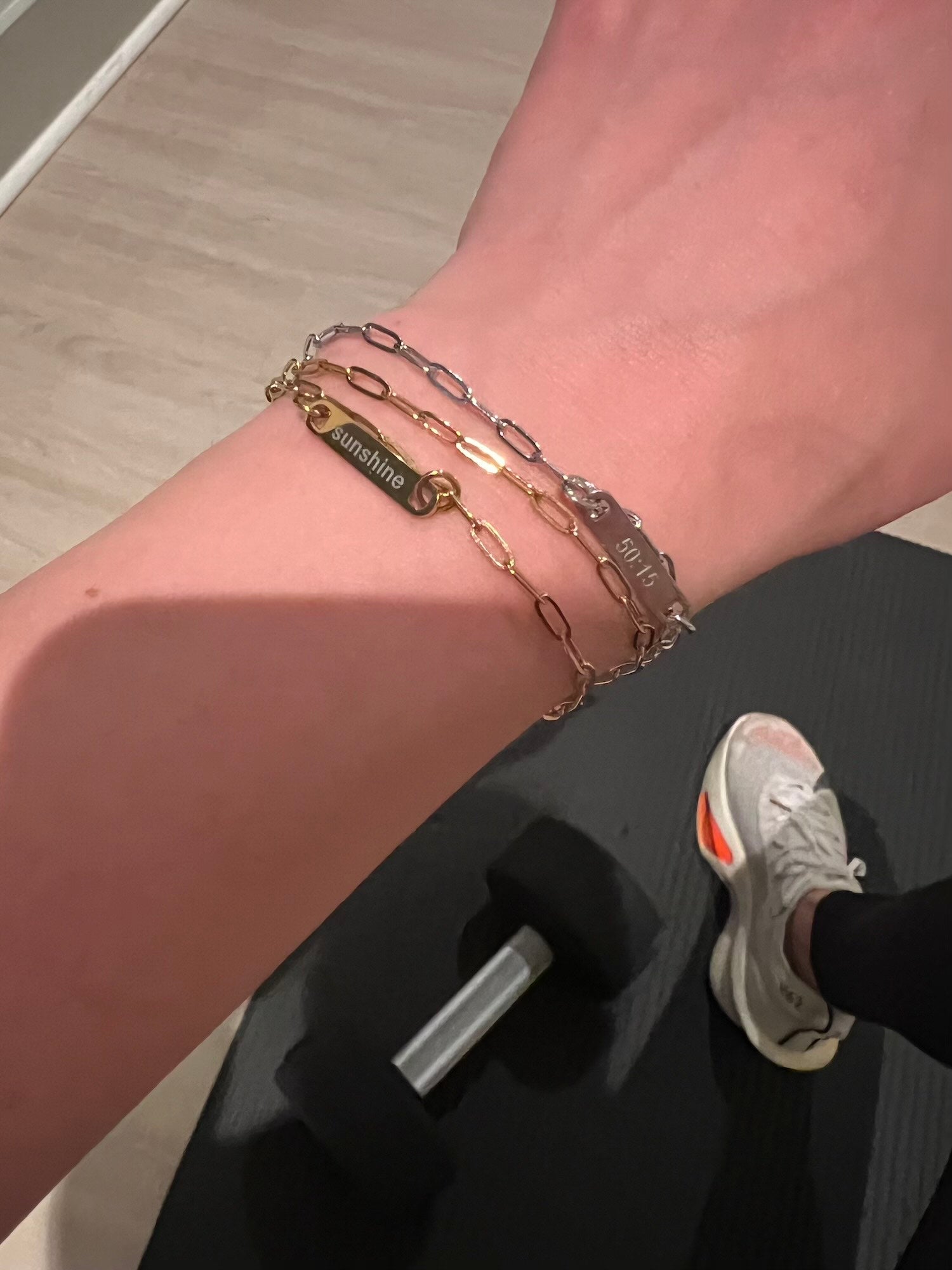 Two bracelets on a wrist in foreground. Generic gym lifestyle image in background of person wearing sneaker on yoga mat with dumbbell on neutral wood floor. Bracelets are paperclip chain with an engraved charm. Model wearing one gold and one silver- each sold separately. Model also wearing third bracelet gold without charm for style.
